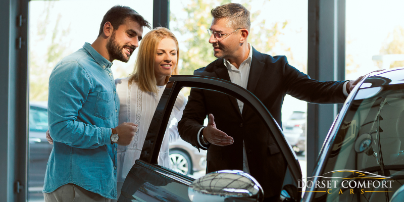 An older male driver inviting a couple of passengers to enter the car with an open door.