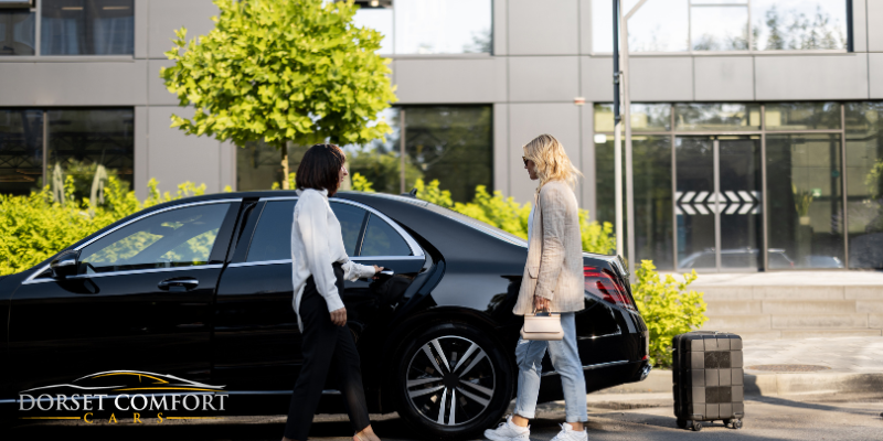 Female driver opening rear left doors to a passenger with a suitcase waiting behind the car to be loaded into the boot.